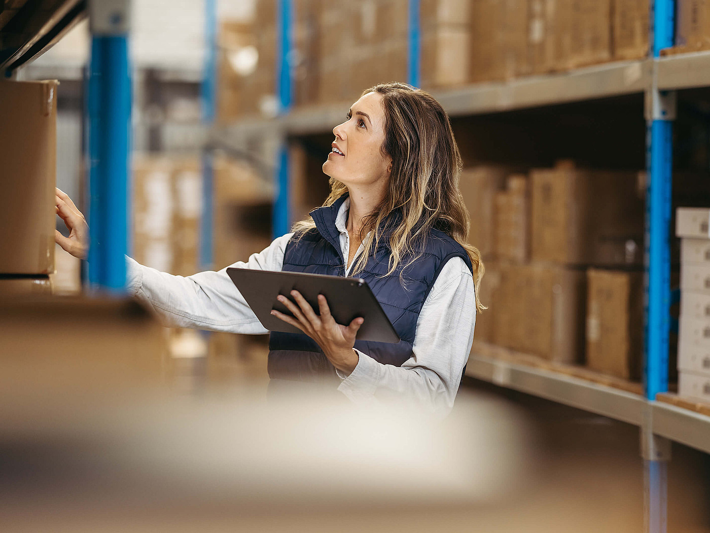 Mitarbeiterin im Lager prüft Kartons und arbeitet mit Tablet zwischen Regalen