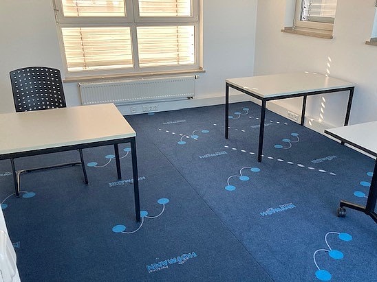 Heller Schulungsraum mit blauem Teppich und vereinzelten Tischen vor großen Fenstern.