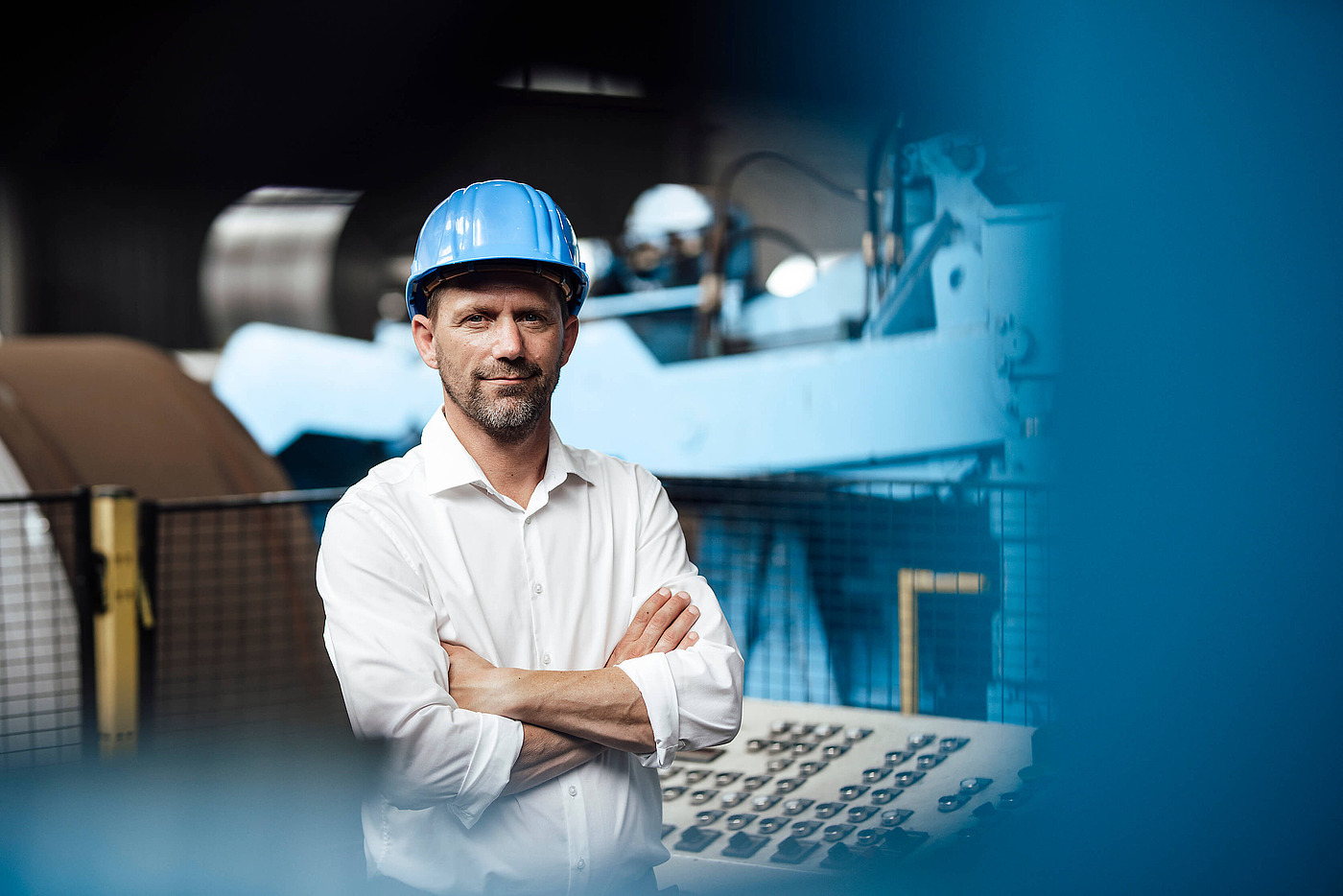 Mitarbeiter mit blauem Schutzhelm und verschränkten Armen in Industrieumgebung
